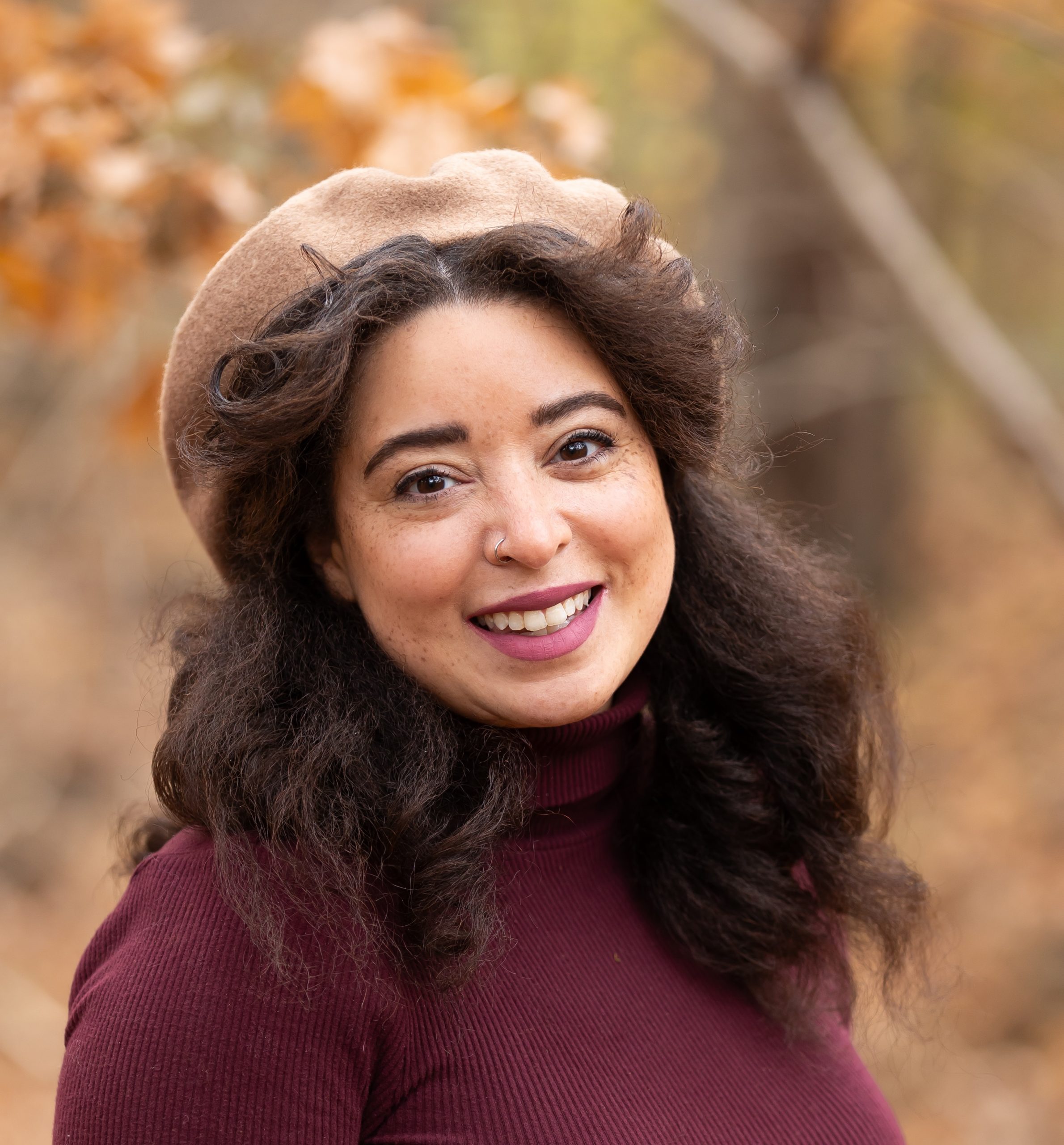 A photo of Nicole Garner Meeker. She is wearing a plum-colored turtleneck, a tan beret, and is standing in front of a few trees in autumn.